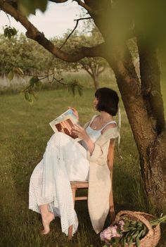 a woman sitting in a chair under a tree reading a book while holding a basket