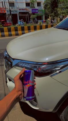 a person holding a can of red bull next to a white car