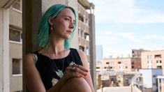 a woman with green hair sitting against a wall in front of a cityscape