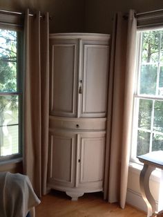 a corner cabinet in the corner of a room with two windows and a table next to it