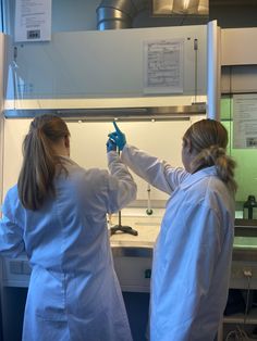 two women in white lab coats are doing something with one hand and the other is pointing