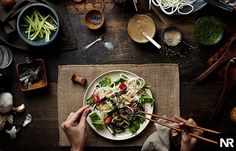 two hands holding chopsticks over a plate of food on a table with other dishes and utensils