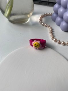 a pink ring with a smiley face on it sitting next to beads and a vase