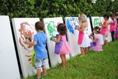 several children are painting on the side of white boards in front of a hedge wall