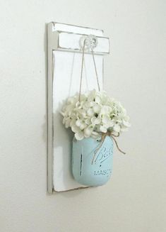 a blue mason jar with white flowers hanging from the side of a wall mounted mirror