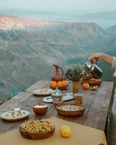 a woman is pouring tea at an outdoor table with food on it and mountains in the background