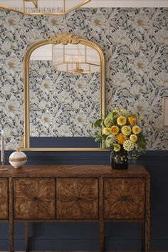 a table with flowers on it in front of a large mirror and wallpapered walls