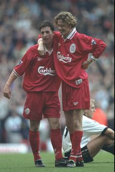 two men in red soccer uniforms standing next to each other on a field with people watching