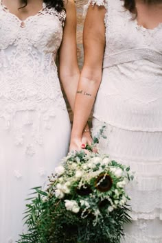 two women in white dresses holding hands with flowers and greenery on the bouquets