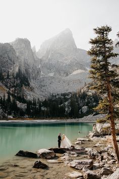 a bride and groom are standing on rocks near the water in front of some mountains