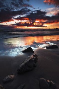 the sun is setting over the ocean with rocks in the foreground and clouds in the background