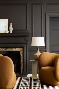 a living room with two chairs and a table in front of a fire place that has a lamp on it