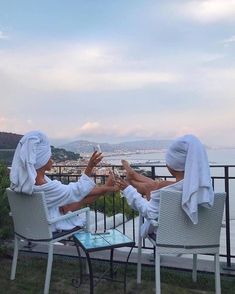 two women in white robes sitting on chairs and taking pictures with their cell phones while overlooking the ocean