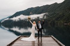 a bride and groom standing on a dock in front of the water with their arms up