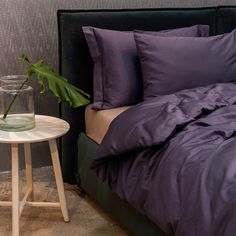 a bed with purple sheets and pillows next to a small table with a plant in it