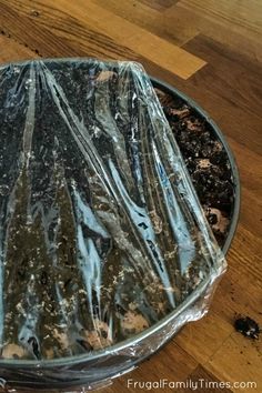 a glass bowl filled with dirt on top of a wooden table covered in plastic wrap