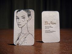 two white business cards sitting on top of a wooden table with a drawing of a woman's face