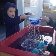 two children are playing in an outdoor water play area, one is pouring water from a container into the other