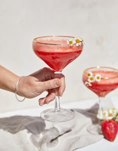 a hand holding a wine glass filled with red liquid and topped with daisies next to two other glasses