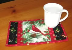 a mug mat with a cardinal and pine cones on it next to a white cup