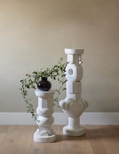 two white vases sitting on top of each other in front of a wall with a plant