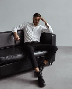 a man sitting on top of a black couch in front of a white wall with his hands behind his head