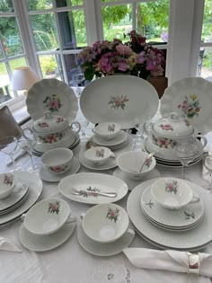 a table topped with lots of white plates and bowls filled with flowers next to a window