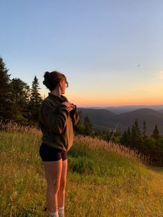 a woman standing on top of a grass covered hillside next to trees and mountains at sunset