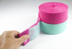 a hand is holding a roll of pink and green tape next to a roll of white paper