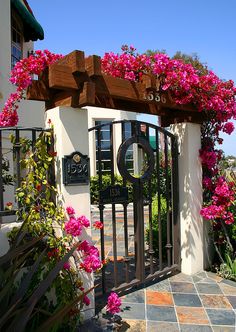 an iron gate with pink flowers growing over it and the entrance to a house in the background