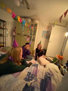 three women in party hats sitting on a bed with balloons and streamers hanging from the ceiling