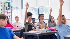 a group of children sitting at desks raising their hands in the air with one hand up