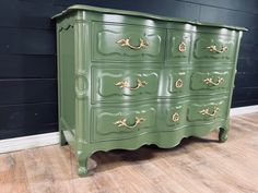 a green dresser with gold handles on it's drawers and wood flooring in front of a black wall