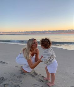 a woman kneeling down next to a baby on top of a sandy beach