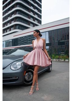 a woman standing next to a car in front of a tall building wearing a pink dress