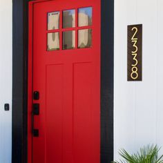 a red front door with black trim on a white wall and potted plant next to it