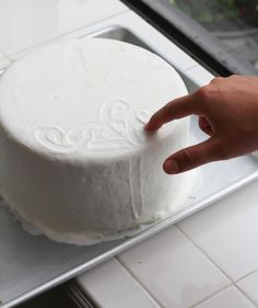 a hand is pointing at a white cake on a metal tray in front of a window