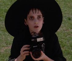 a woman wearing a black hat and holding a camera in front of her face while sitting on the grass