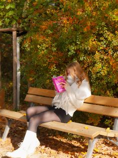 a woman is sitting on a bench reading a book in the fall leaves with her legs crossed