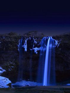 a waterfall is lit up at night with snow on the ground and trees in the foreground