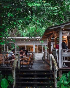 people are sitting at wooden tables on the porch in front of a tree covered building