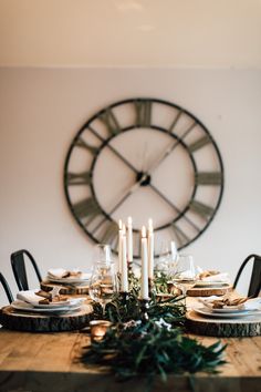 a wooden table topped with plates and candles next to a large clock on the wall