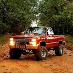 a red pickup truck driving down a dirt road in the middle of some trees and bushes