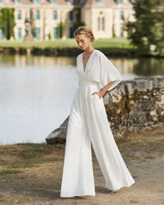 a woman standing in front of a body of water wearing a white jumpsuit and matching heels