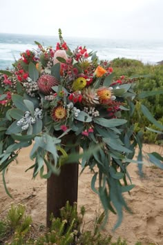 an arrangement of flowers and greenery in a vase on the sand near the ocean