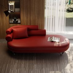 a red couch sitting in front of a window next to a book on top of a table