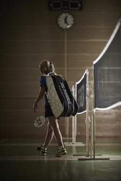 a woman with a back pack and tennis racket walks towards the net in an indoor court