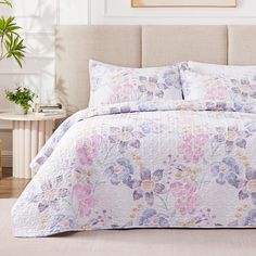 a bed covered in a white and purple floral comforter with pillows on top of it