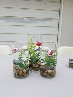 three clear vases filled with plants and rocks on a white tablecloth covered table