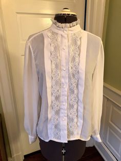 "This is an easy care yet professional blouse. This blouse is made of 100% polyester crepe and lace. There are no shoulder pads. It features a band collar with lace, front pearl button closure with vertical lace and a one inch tuck on either side. There are long sleeves with button cuffs, and a front pearl button closure. This garment is clean with no stains, snags, or odors. Measurements to guide you: Bust: 38\" Waist: 40\" Sleeve: 24\" Length from top of shoulder to hem: 26\" Decade: 70s Fabri Fitted Lace Trim Top For Work, Formal Lace Blouse With Ruffles, Formal Blouse With Lace Trim And Ruffled Collar, Elegant Blouse With Lace Trim And Ruffled Collar, Lace Blouse With Lace Cuffs For Work, Elegant Blouse With Lace Cuffs For Daywear, Elegant Lace Work Blouse For Daywear, Elegant Fitted Tops With Ruffled Collar, Feminine Blouse With Ruffled Collar For Formal Occasions